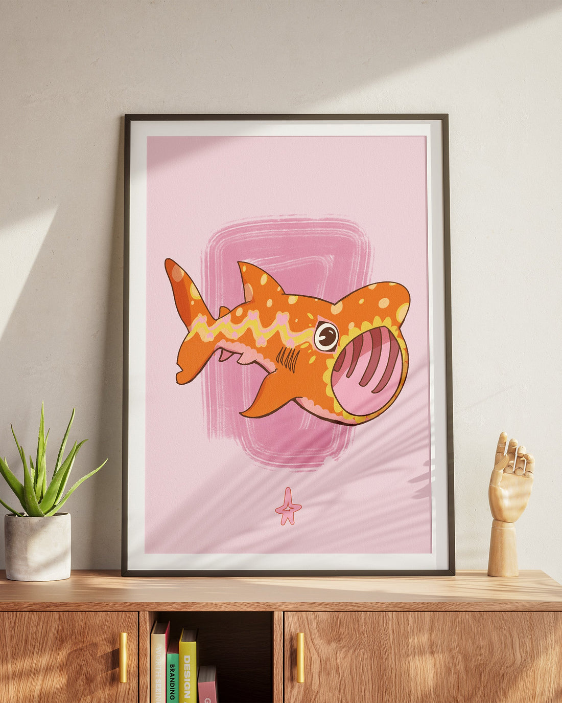 Framed illustration of a cartoon shark on a pink background, displayed on a wooden cabinet.