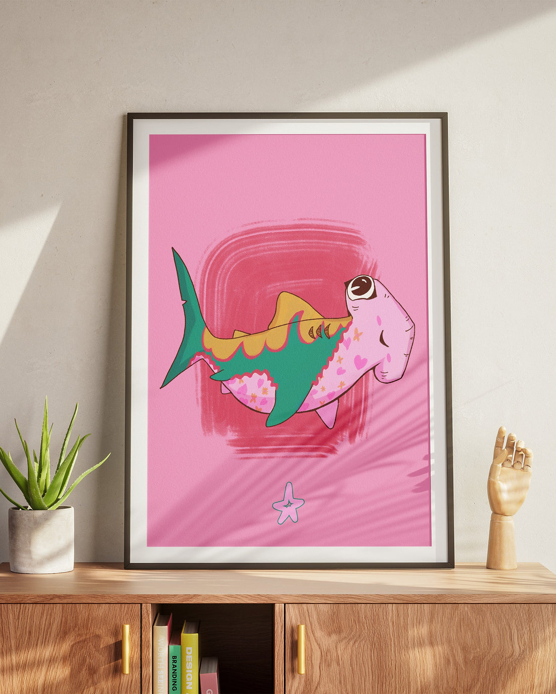 Framed artwork of a pink shark on a pink background, displayed on a wooden cabinet.