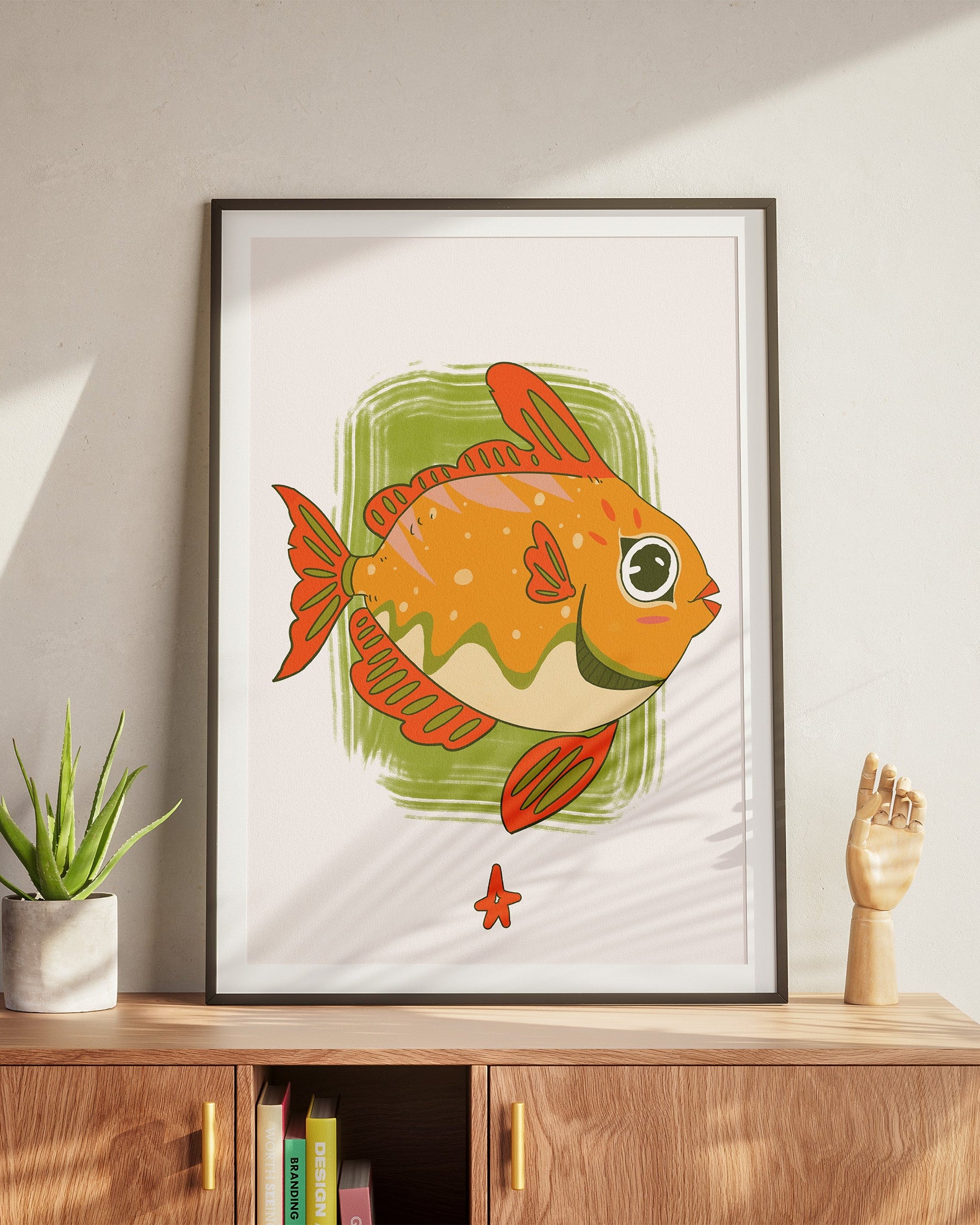 Framed illustration of a goldfish on a wall above a wooden cabinet with a plant and sculpture.