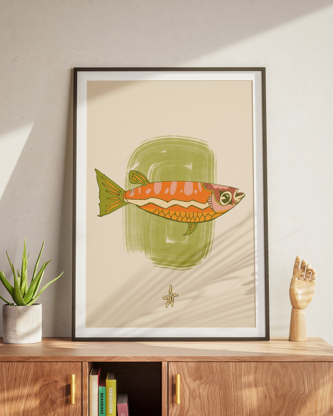 Framed illustration of a fish on a wall above a wooden cabinet with a plant and sculpture.