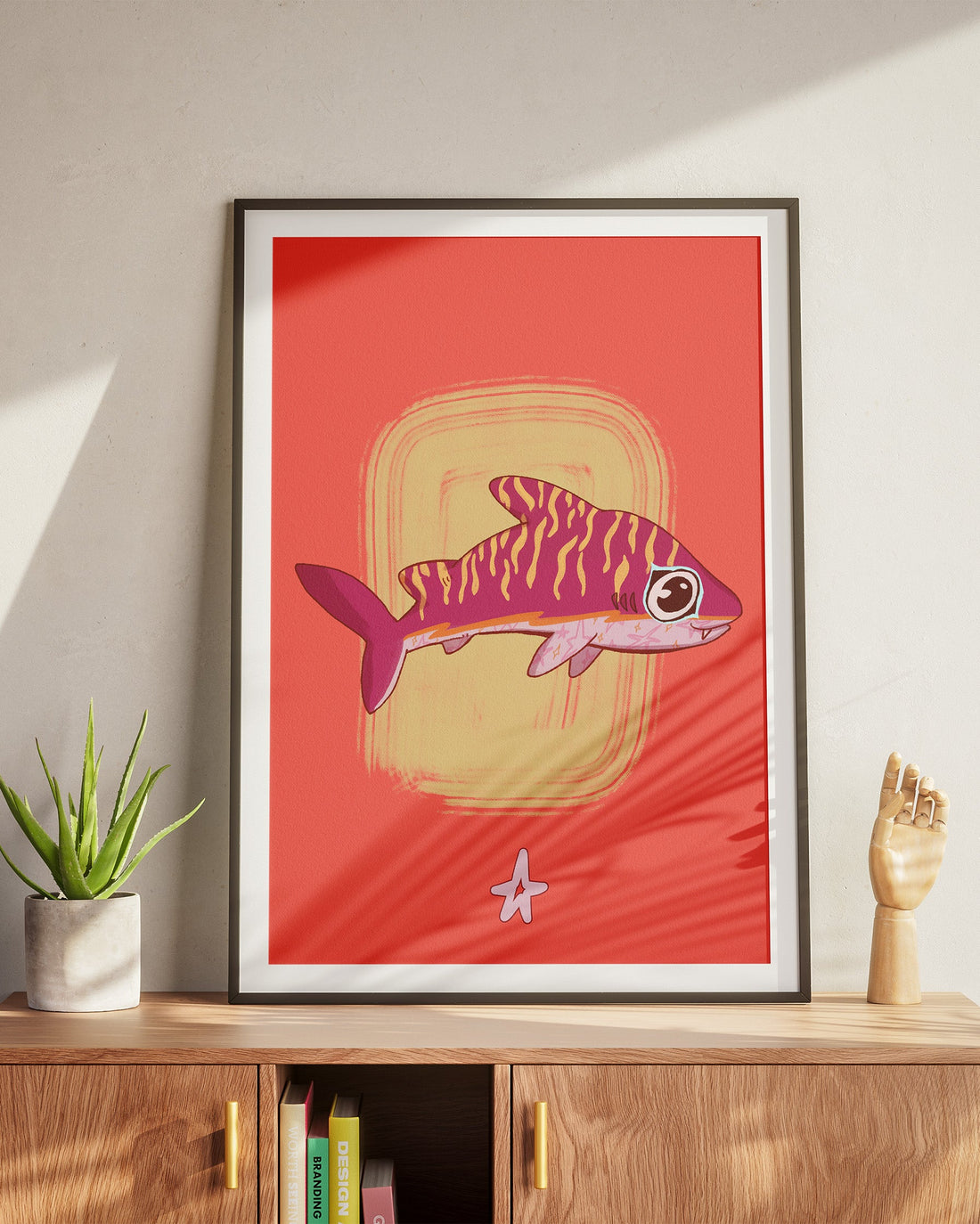 Framed artwork of a fish on a red background in a room with a wooden cabinet and plant.